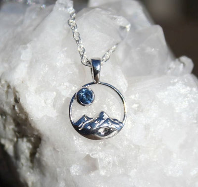 A silver pendant necklace featuring a stylized mountain and moon design with a small blue Montana sapphire, displayed on a textured white crystal cluster.