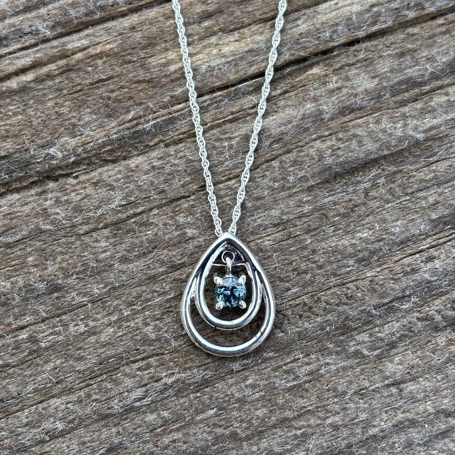 A silver teardrop pendant necklace with a small blue sapphire cluster, displayed on a rustic, weathered wood background.
