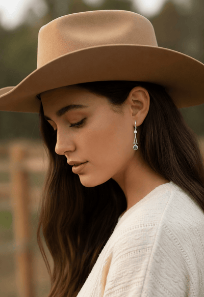 A woman wearing a tan cowboy hat and long drop Montana sapphire earrings with a larger blue sapphire at the bottom.