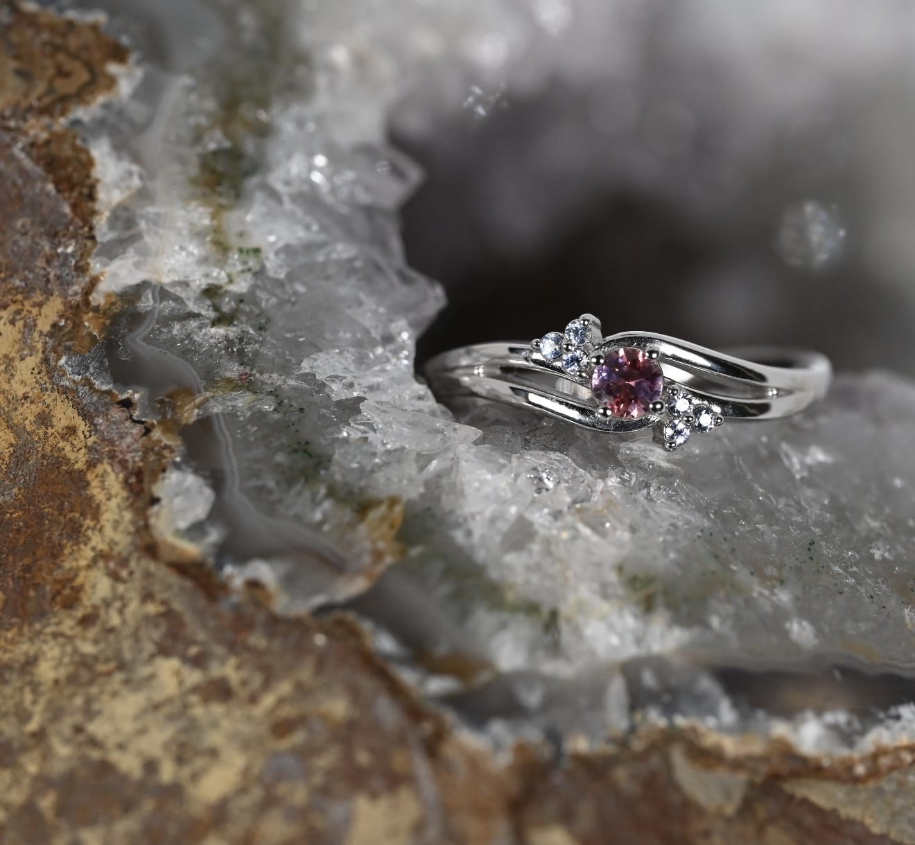 A white gold ring with a central round pink gemstone and small lavender accent stones.