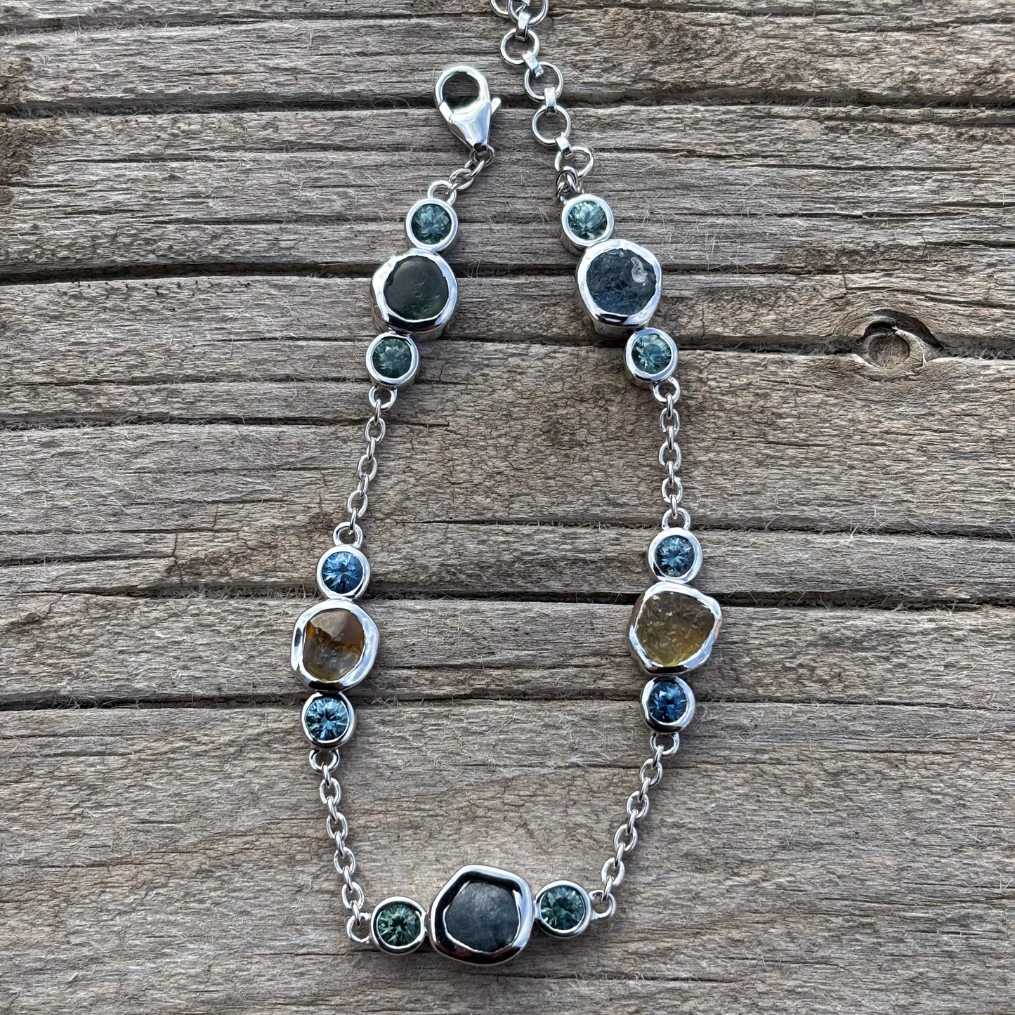 White gold chain style bracelet with cut and uncut sapphires on a wooden surface