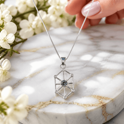 A silver geometric pendant necklace with a central blue gemstone, held by a hand over a white marble slab with white flowers.
