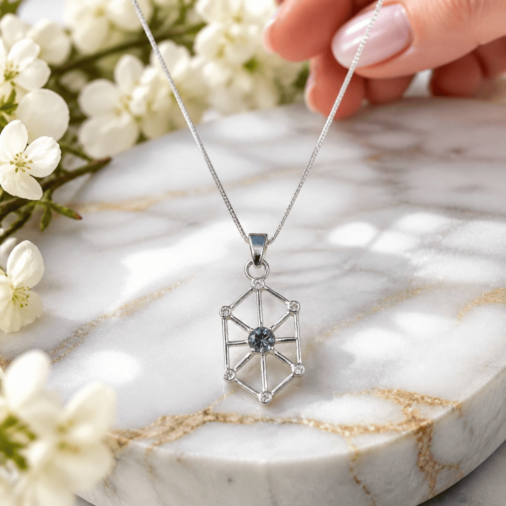A silver geometric pendant necklace with a central blue gemstone, held by a hand over a white marble slab with white flowers.