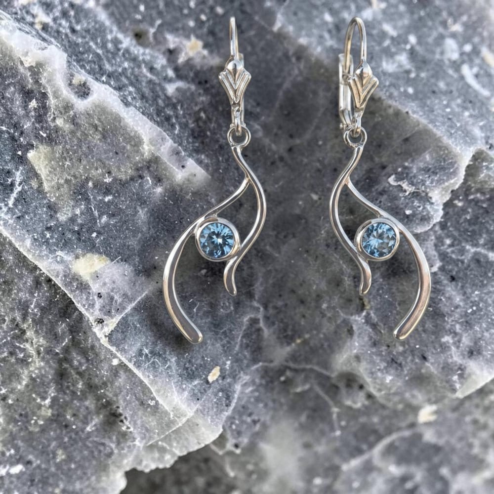 A pair of silver bezel set sapphires in a drop earring style with a curved wave design and a round light blue sapphire.