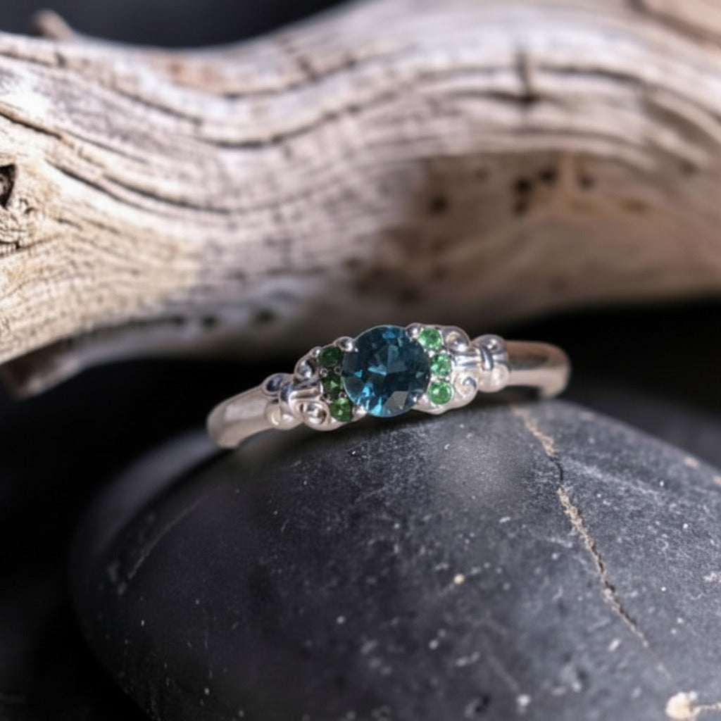 A white gold three-stone ring featuring a central round blue Montana sapphire flanked by six small green tsavorite garnets.
