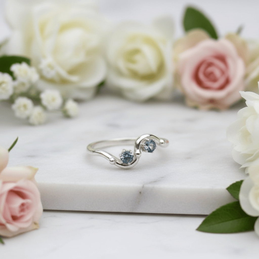 A silver ring with two small round blue sapphires, displayed on a white marble slab surrounded by soft pink and white roses.