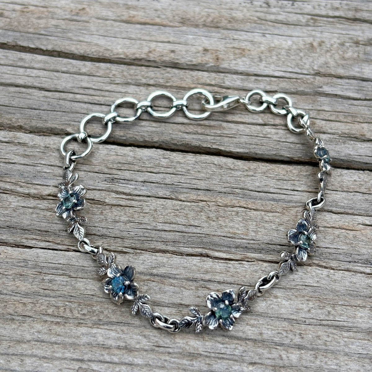 A silver chain bracelet with floral links with blue hued Montana sapphires at the center of each flower.