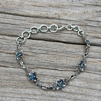 A silver chain bracelet with floral links with blue hued Montana sapphires at the center of each flower.