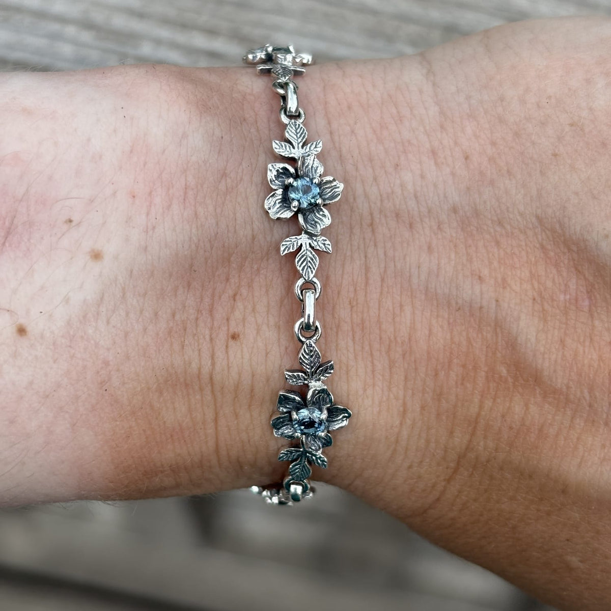 A silver floral chain bracelet with blue Montana sapphires inside the flower details, shown worn on a person's wrist.