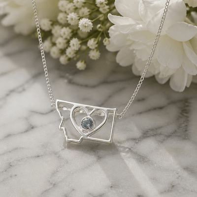 A silver necklace featuring an open Montana state silhouette pendant with a heart-shaped cutout and a round Montana sapphire bezel set in the center. 