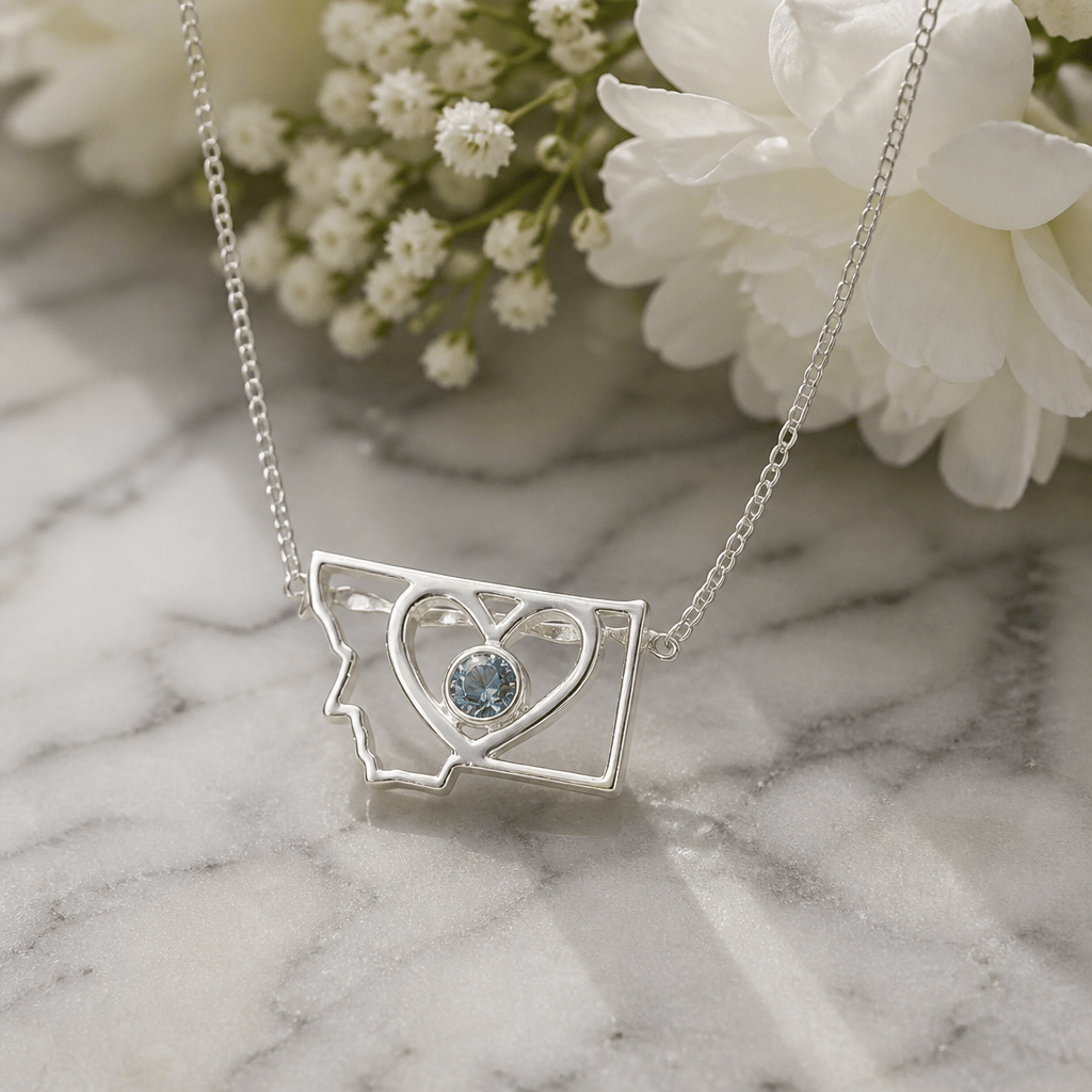 A silver necklace featuring an open Montana state silhouette pendant with a heart-shaped cutout and a round Montana sapphire bezel set in the center. 