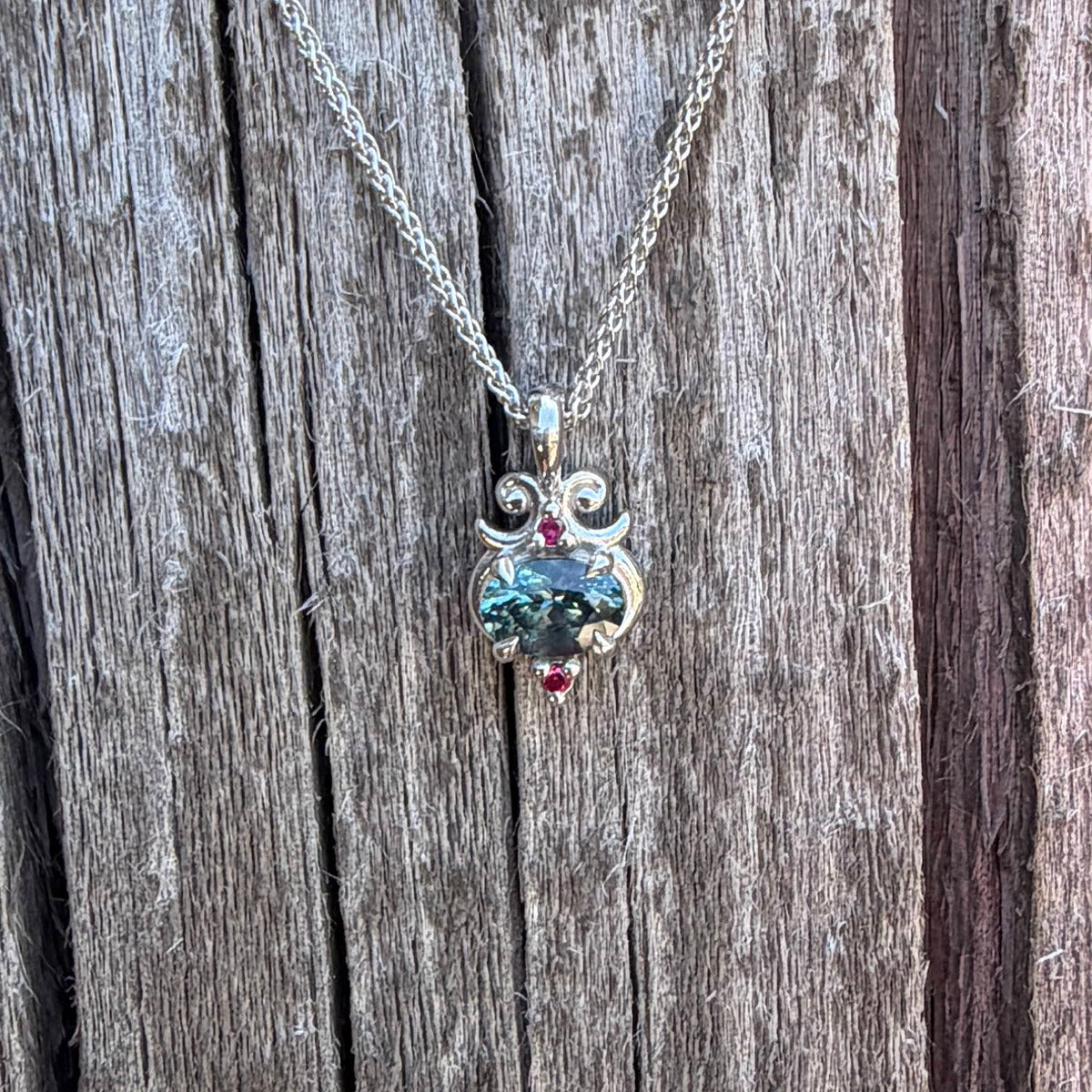 A silver pendant necklace with a teal Montana sapphire and two small ruby accent stones, displayed on a rustic weathered wood background.