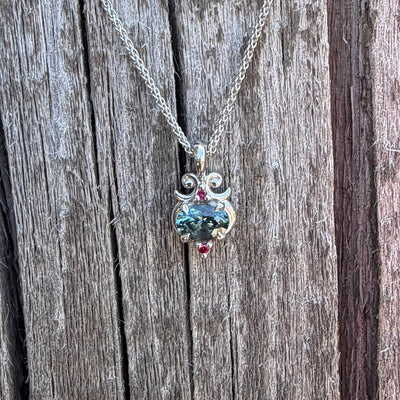 A silver pendant necklace with a teal Montana sapphire and two small ruby accent stones, displayed on a rustic weathered wood background.