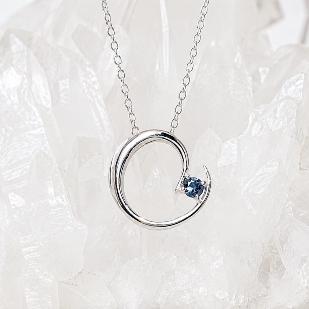A silver necklace with a modern open circular pendant featuring a small round blue Montana sapphire.