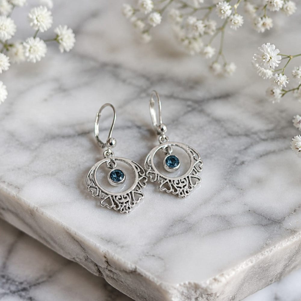 A pair of silver drop earrings with intricate filigree cutouts and a small round blue Montana sapphire gemstone in the center