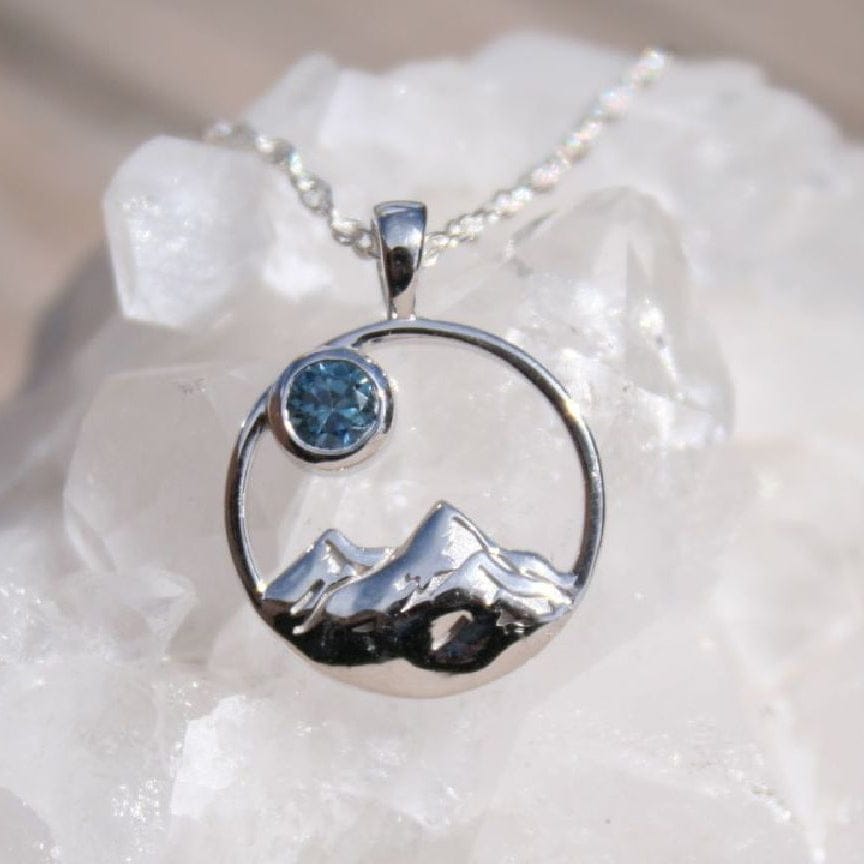 Silver pendant with mountain scene in a round frame.  Has a 3mm bezel set blue Montana sapphire. 