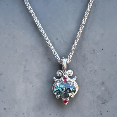 A silver pendant modern style necklace featuring an oval, teal Montana sapphire with 2 small ruby accents. 