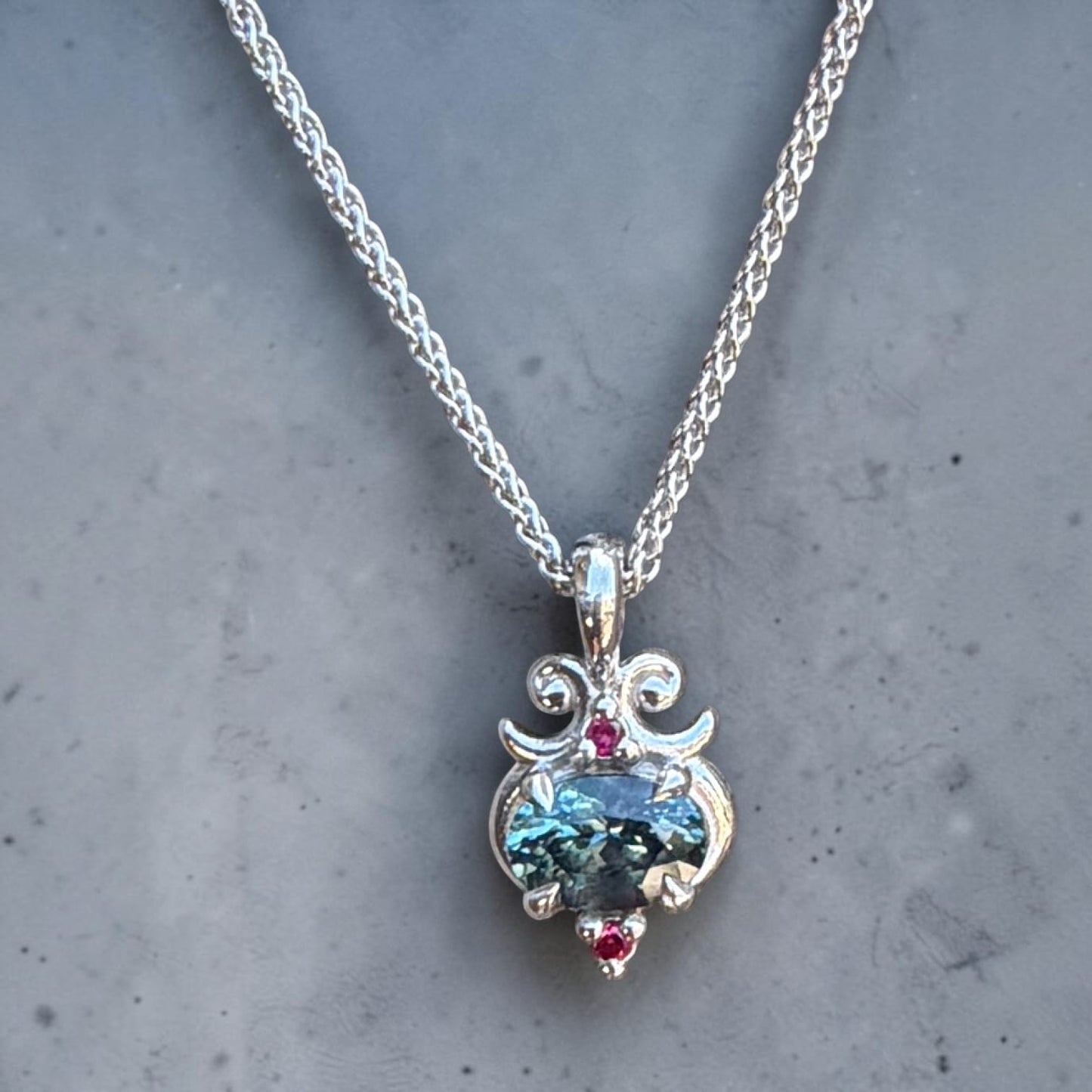 A silver pendant modern style necklace featuring an oval, teal Montana sapphire with 2 small ruby accents. 